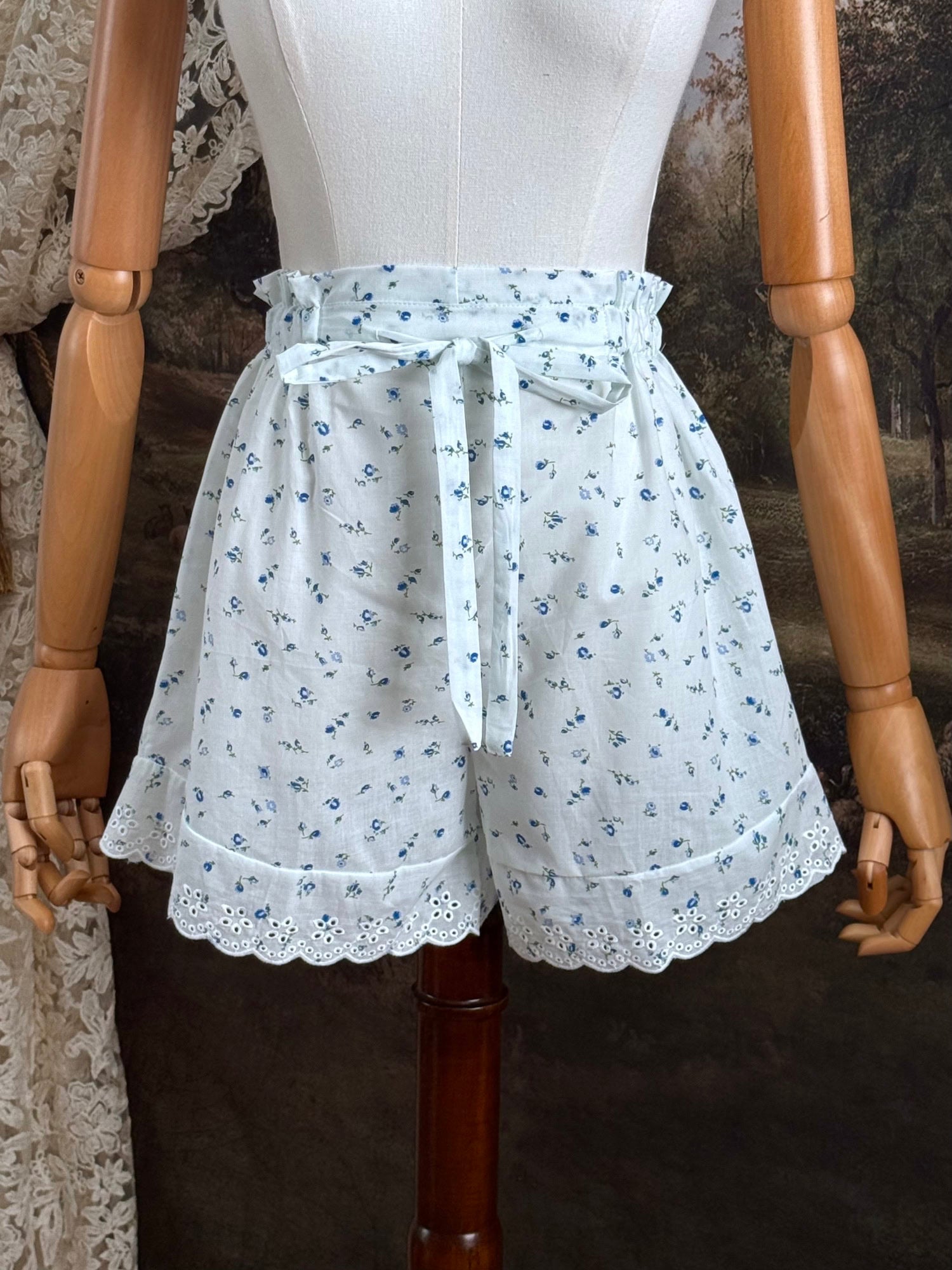 A pair of vintage inspired white and blue floral print cotton bloomer shorts with scalloped lace trim and bow-tied waist drawstring, pictured on a mannequin.