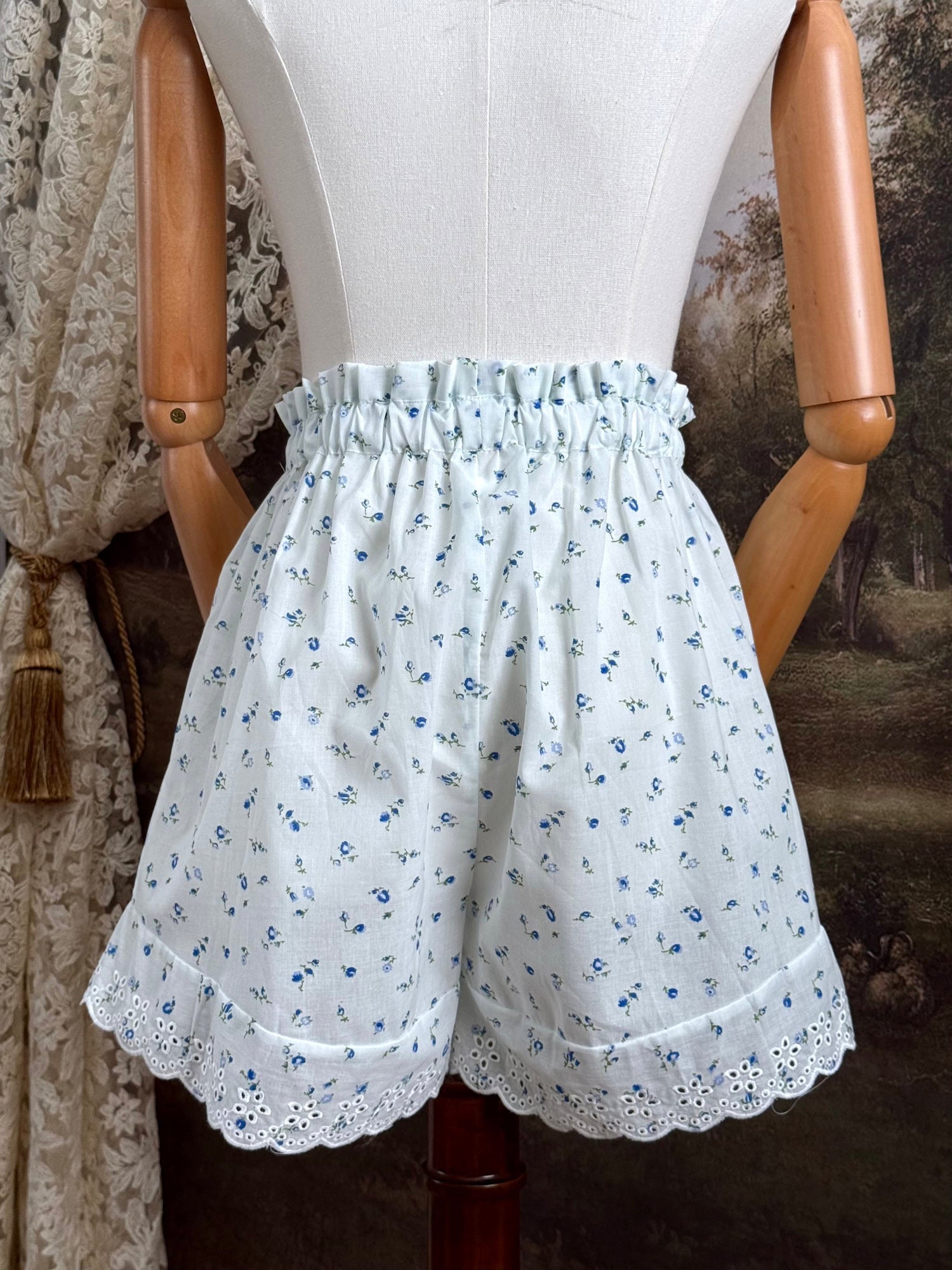 A pair of vintage inspired white and blue floral print cotton bloomer shorts with scalloped lace trim and bow-tied waist drawstring, pictured on a mannequin.