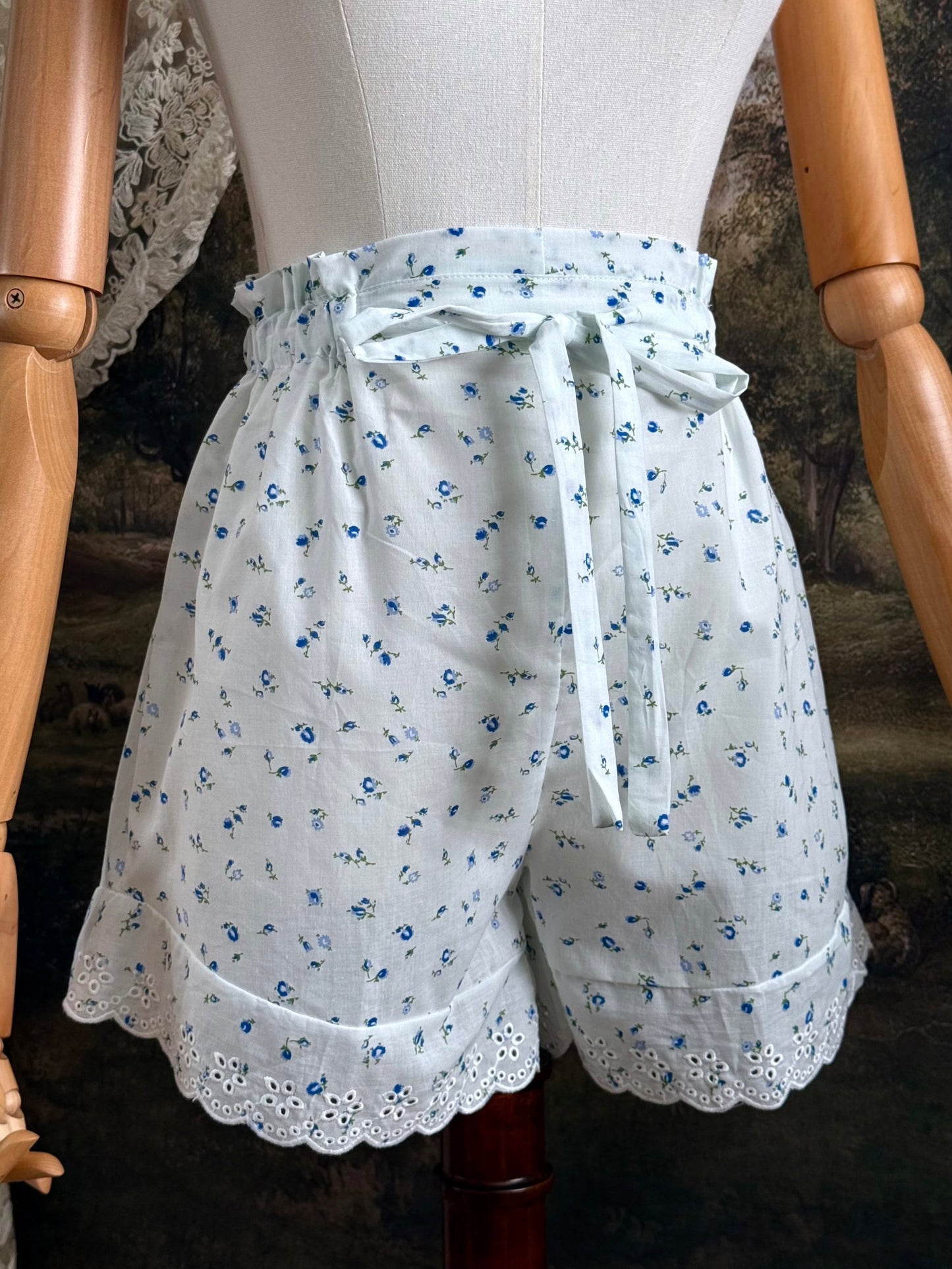 A pair of vintage inspired white and blue floral print cotton bloomer shorts with scalloped lace trim and bow-tied waist drawstring, pictured on a mannequin.