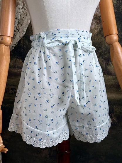 A pair of vintage inspired white and blue floral print cotton bloomer shorts with scalloped lace trim and bow-tied waist drawstring, pictured on a mannequin.