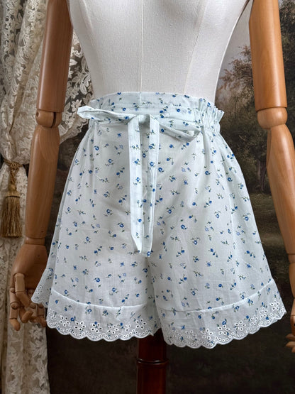 A pair of vintage inspired white and blue floral print cotton bloomer shorts with scalloped lace trim and bow-tied waist drawstring, pictured on a mannequin.