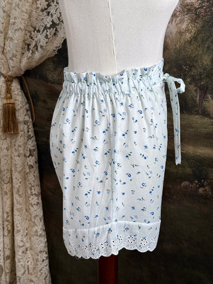 A pair of vintage inspired white and blue floral print cotton bloomer shorts with scalloped lace trim and bow-tied waist drawstring, pictured on a mannequin.