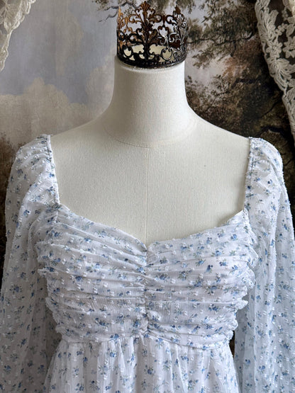A historically Inspired regency style empire waist mini dress with ruffle hem, ruched bodice, and tulip sleeves made of white and blue floral chiffon. Pictured on a mannequin in front of an art history backdrop.