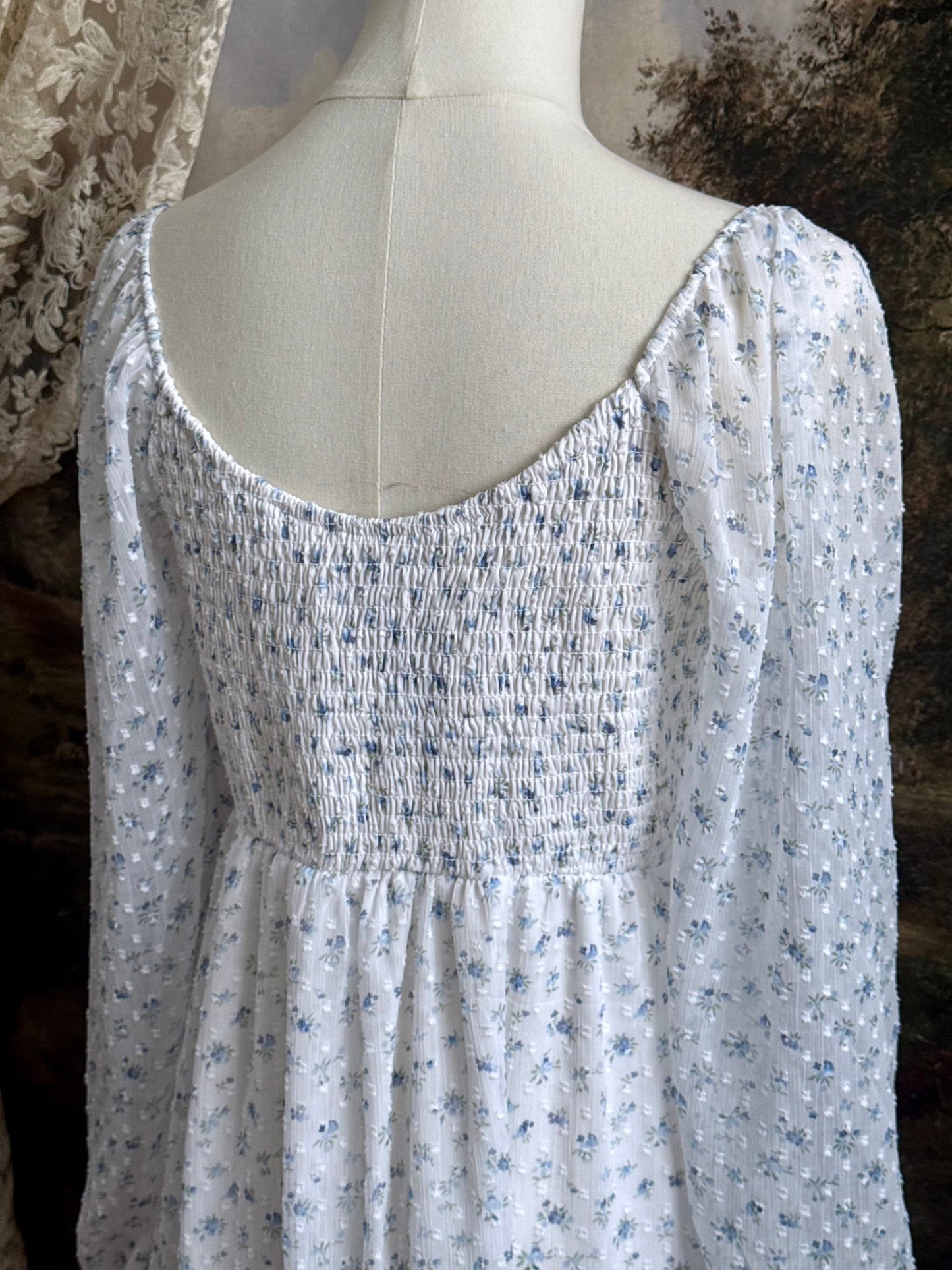 A historically Inspired regency style empire waist mini dress with ruffle hem, ruched bodice, and tulip sleeves made of white and blue floral chiffon. Pictured on a mannequin in front of an art history backdrop.
