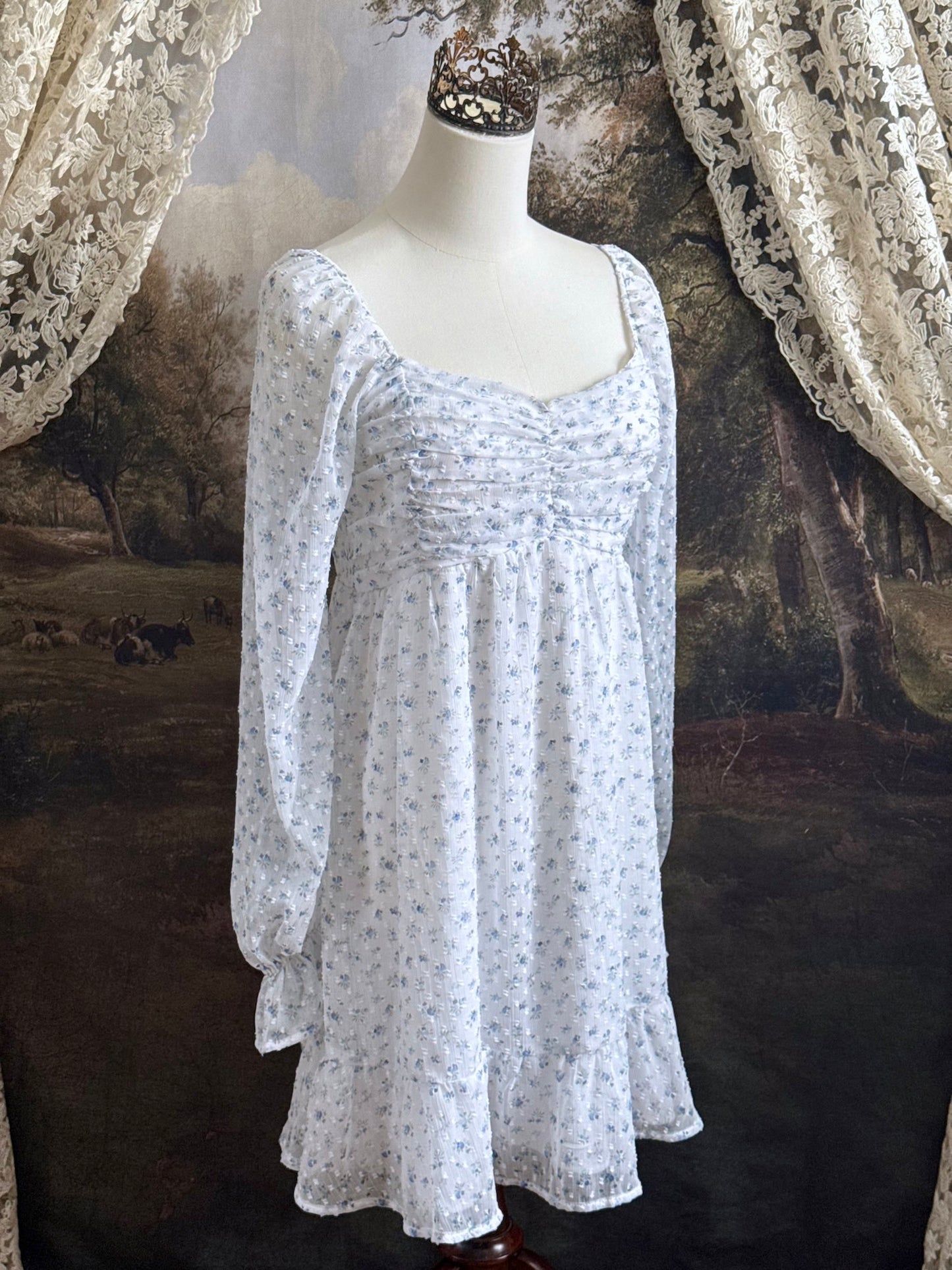 A historically Inspired regency style empire waist mini dress with ruffle hem, ruched bodice, and tulip sleeves made of white and blue floral chiffon. Pictured on a mannequin in front of an art history backdrop.