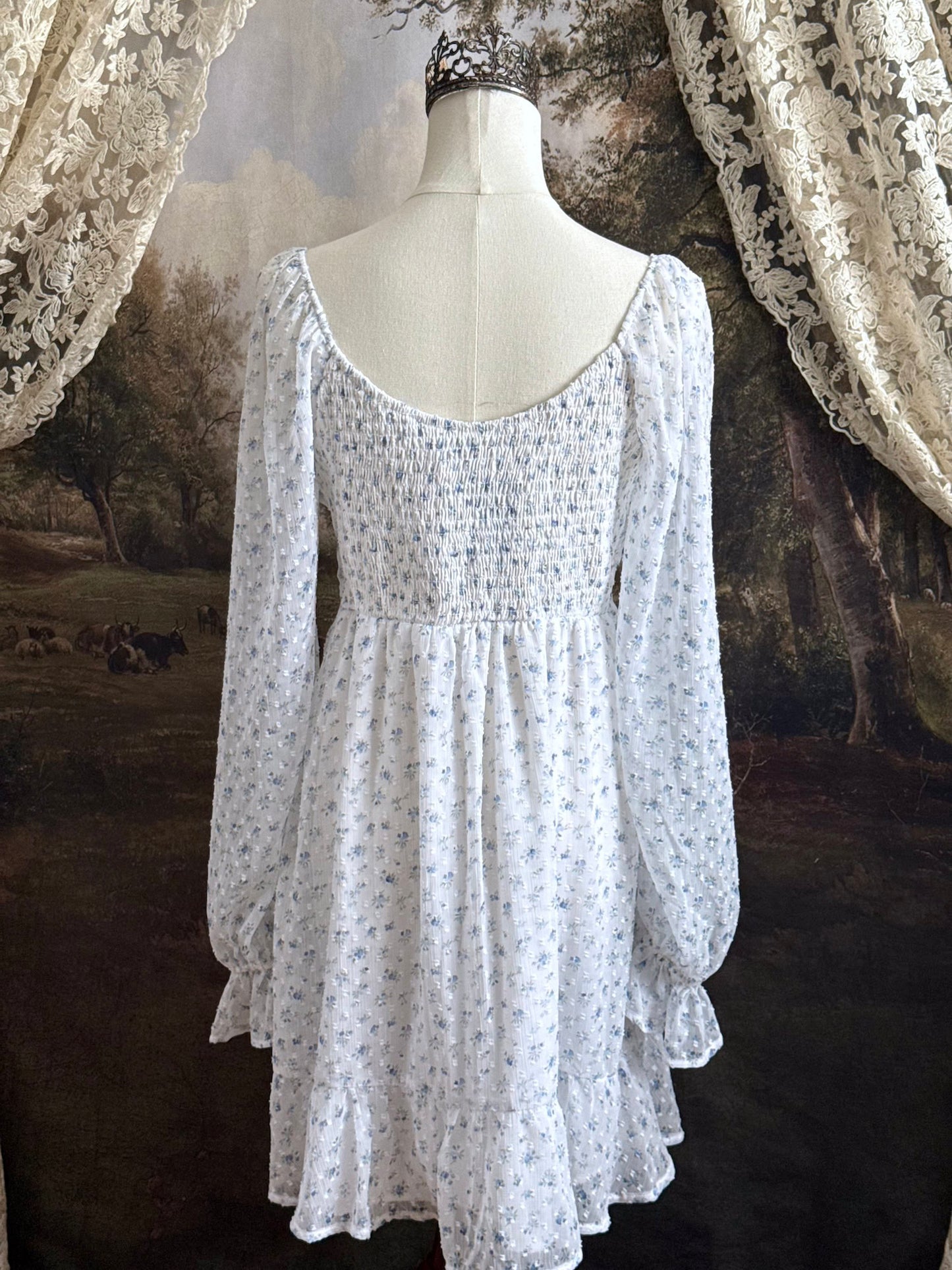 A historically Inspired regency style empire waist mini dress with ruffle hem, ruched bodice, and tulip sleeves made of white and blue floral chiffon. Pictured on a mannequin in front of an art history backdrop.