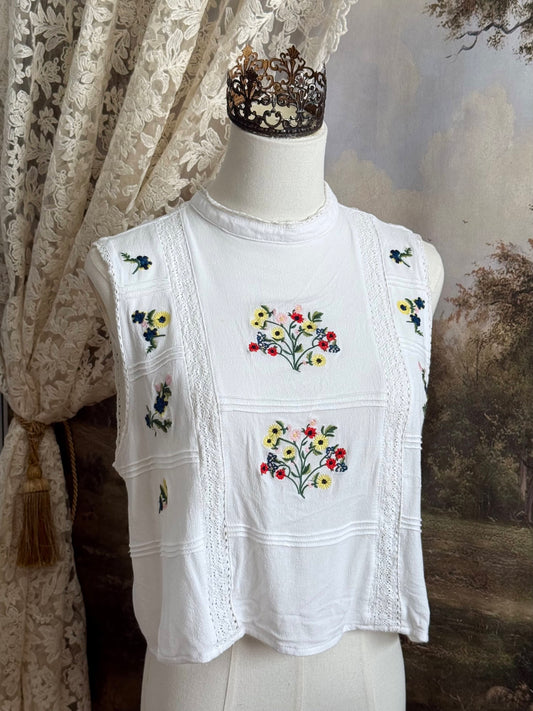 White edwardian-inspired sleeveless tank top blouse with floral embroidery and lace trim details, pictured on a mannequin against an art history background.