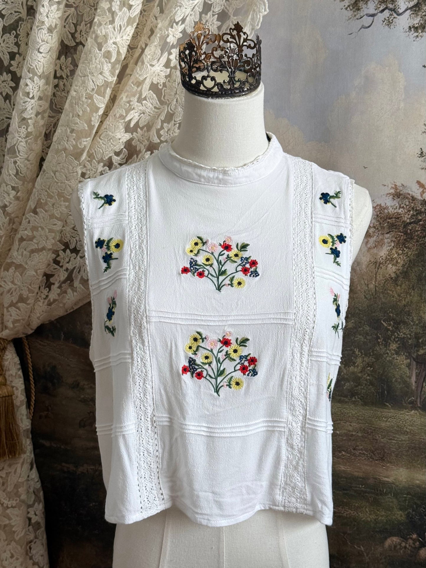 White edwardian-inspired sleeveless tank top blouse with floral embroidery and lace trim details, pictured on a mannequin against an art history background.
