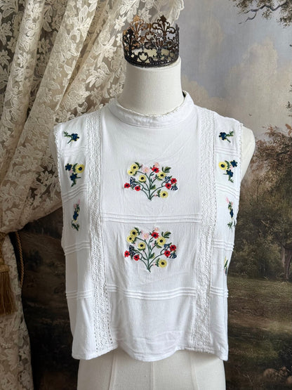 White edwardian-inspired sleeveless tank top blouse with floral embroidery and lace trim details, pictured on a mannequin against an art history background.