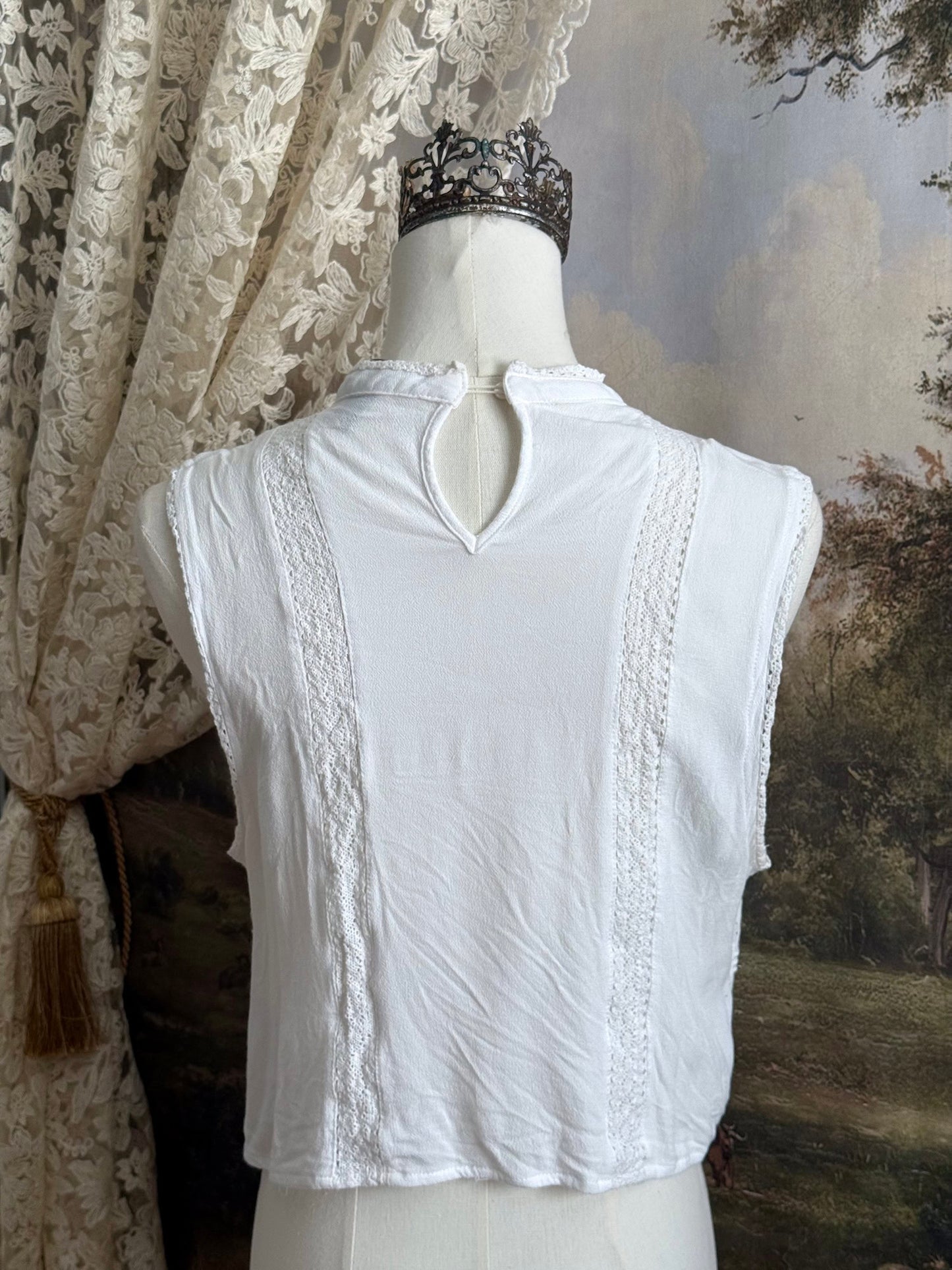 White edwardian-inspired sleeveless tank top blouse with floral embroidery and lace trim details, pictured on a mannequin against an art history background.