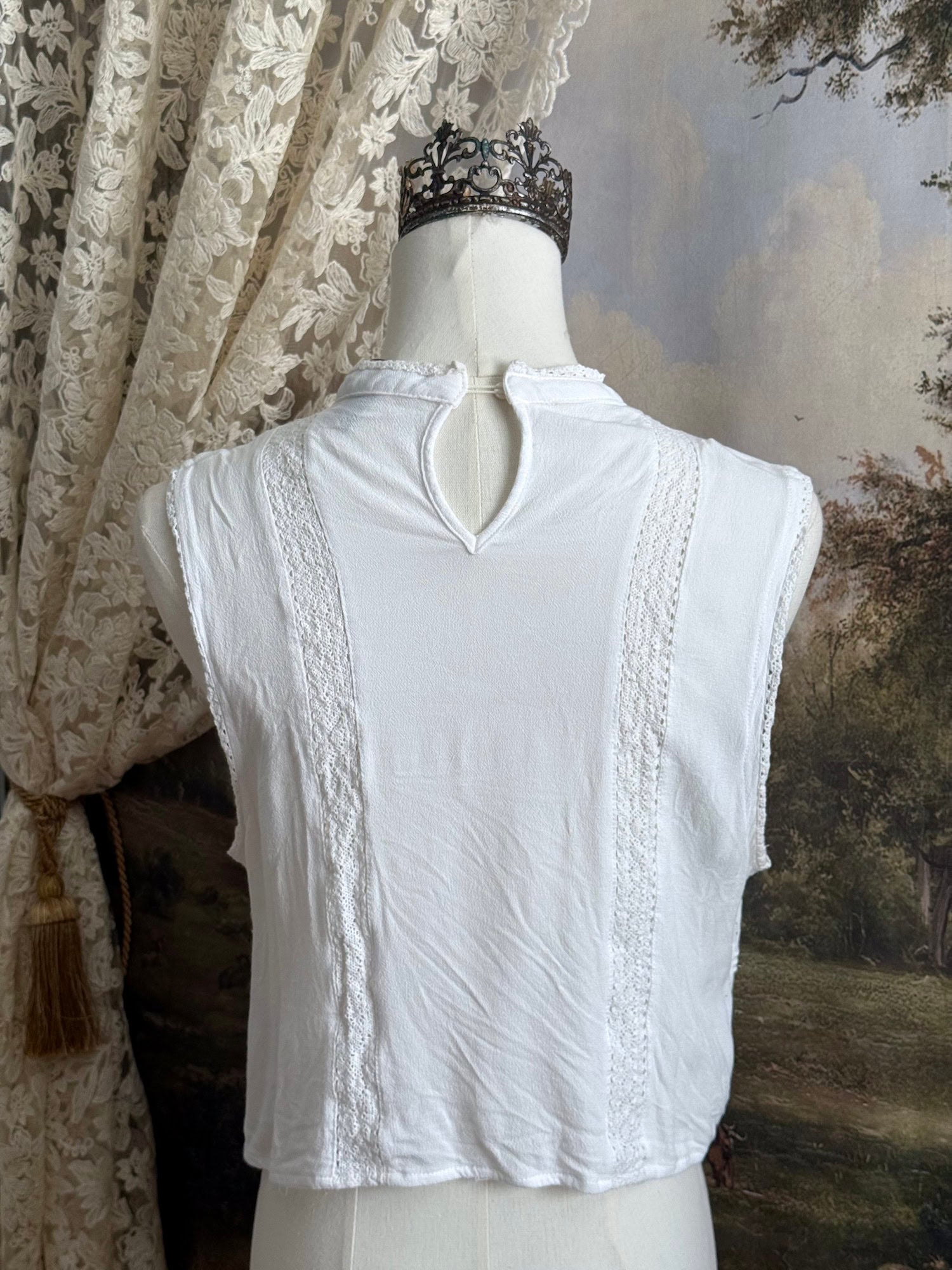 White edwardian-inspired sleeveless tank top blouse with floral embroidery and lace trim details, pictured on a mannequin against an art history background.