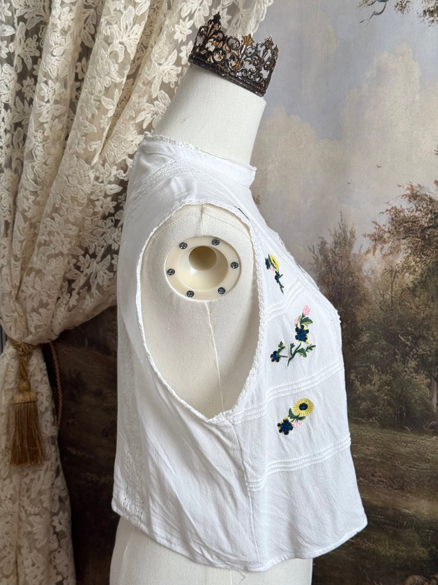 White edwardian-inspired sleeveless tank top blouse with floral embroidery and lace trim details, pictured on a mannequin against an art history background.