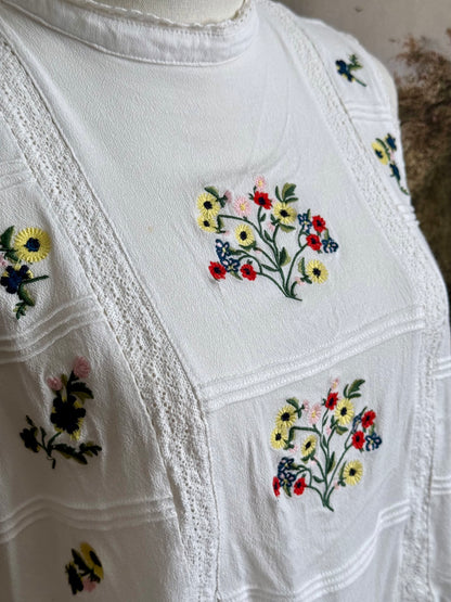 White edwardian-inspired sleeveless tank top blouse with floral embroidery and lace trim details, pictured on a mannequin against an art history background.