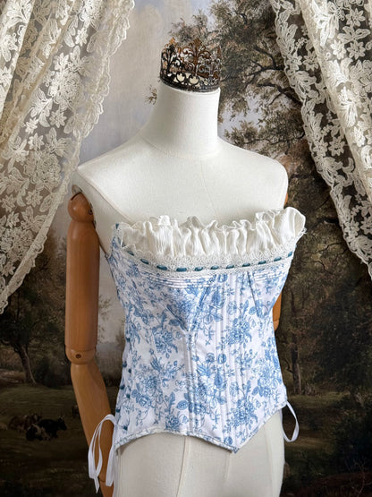 A historically-inspired vintage style overbust bustier corset in white and blue floral toile print, with a pointed basque waist, velvet lacing ties, lace trim, and a ruched bust detail, pictured on a mannequin.