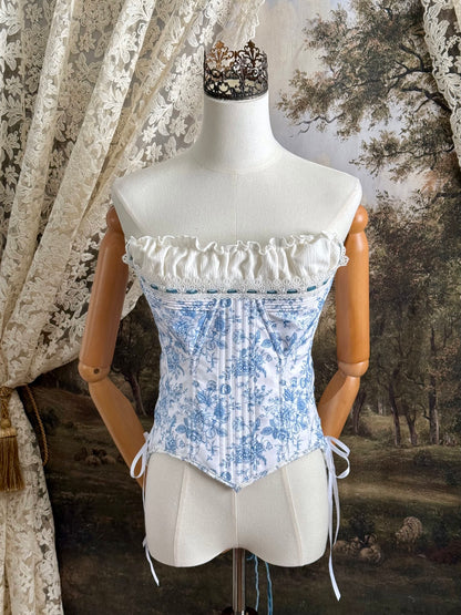 A historically-inspired vintage style overbust bustier corset in white and blue floral toile print, with a pointed basque waist, velvet lacing ties, lace trim, and a ruched bust detail, pictured on a mannequin.
