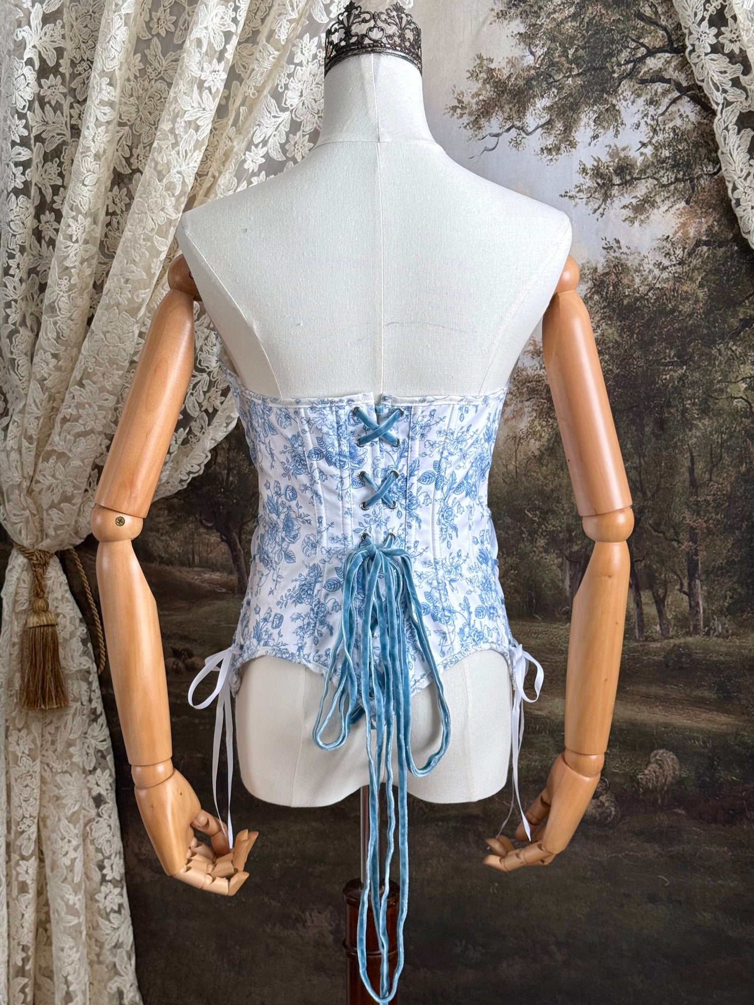 A historically-inspired vintage style overbust bustier corset in white and blue floral toile print, with a pointed basque waist, velvet lacing ties, lace trim, and a ruched bust detail, pictured on a mannequin. Back details.