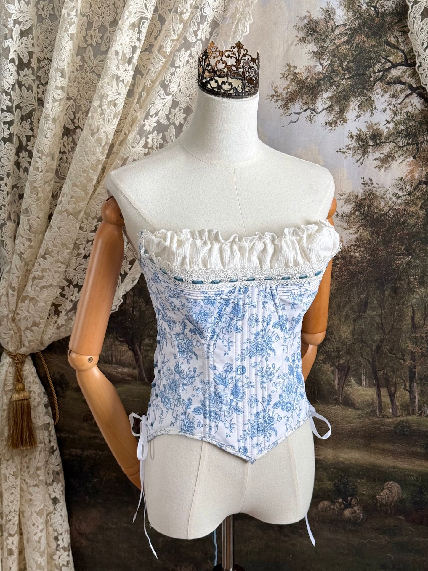 A historically-inspired vintage style overbust bustier corset in white and blue floral toile print, with a pointed basque waist, velvet lacing ties, lace trim, and a ruched bust detail, pictured on a mannequin.