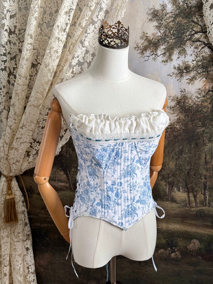 A historically-inspired vintage style overbust bustier corset in white and blue floral toile print, with a pointed basque waist, velvet lacing ties, lace trim, and a ruched bust detail, pictured on a mannequin.