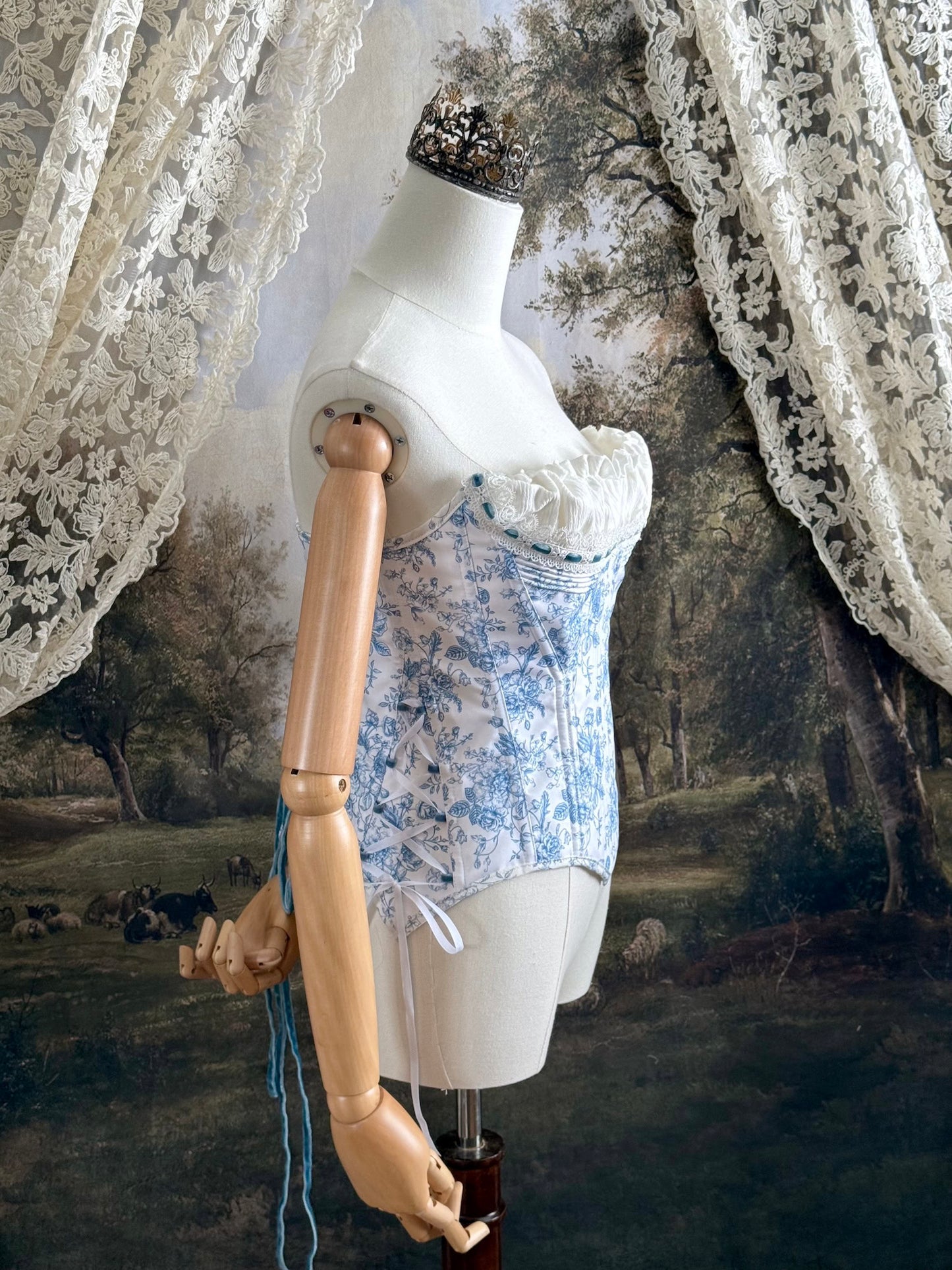 A historically-inspired vintage style overbust bustier corset in white and blue floral toile print, with a pointed basque waist, velvet lacing ties, lace trim, and a ruched bust detail, pictured on a mannequin.