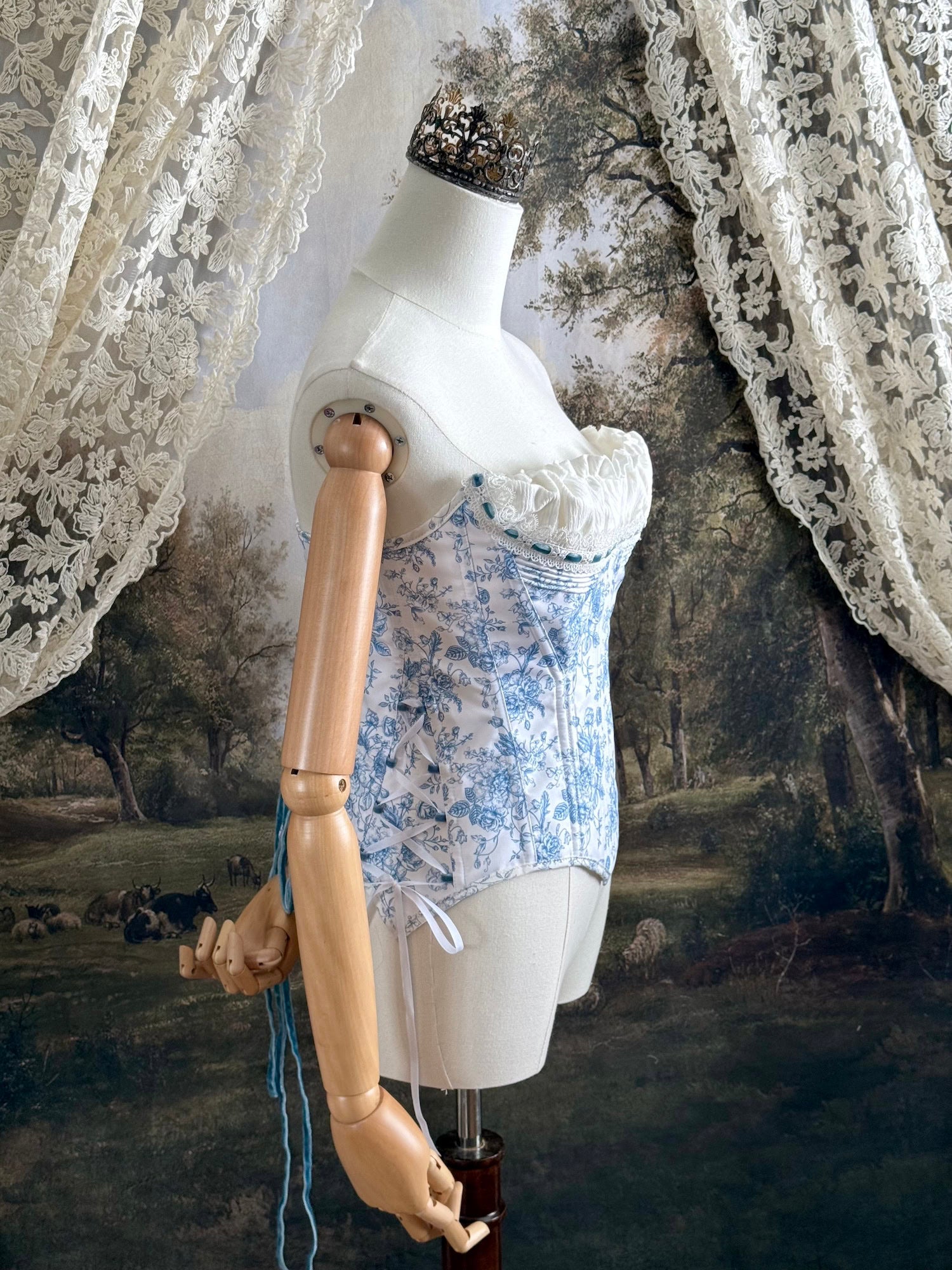 A historically-inspired vintage style overbust bustier corset in white and blue floral toile print, with a pointed basque waist, velvet lacing ties, lace trim, and a ruched bust detail, pictured on a mannequin.