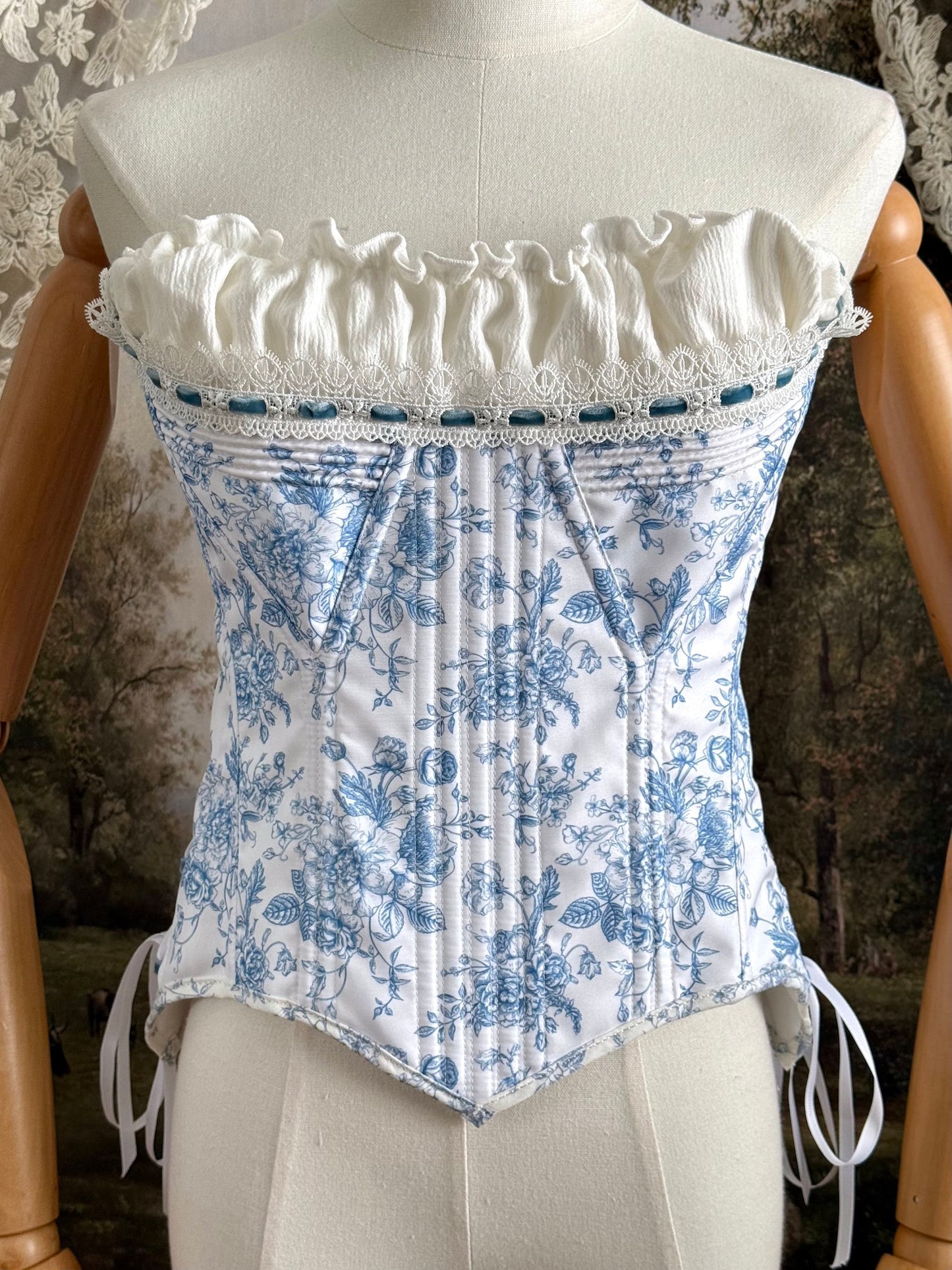 A historically-inspired vintage style overbust bustier corset in white and blue floral toile print, with a pointed basque waist, velvet lacing ties, lace trim, and a ruched bust detail, pictured on a mannequin.
