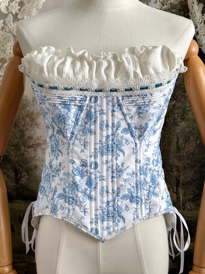 A historically-inspired vintage style overbust bustier corset in white and blue floral toile print, with a pointed basque waist, velvet lacing ties, lace trim, and a ruched bust detail, pictured on a mannequin.