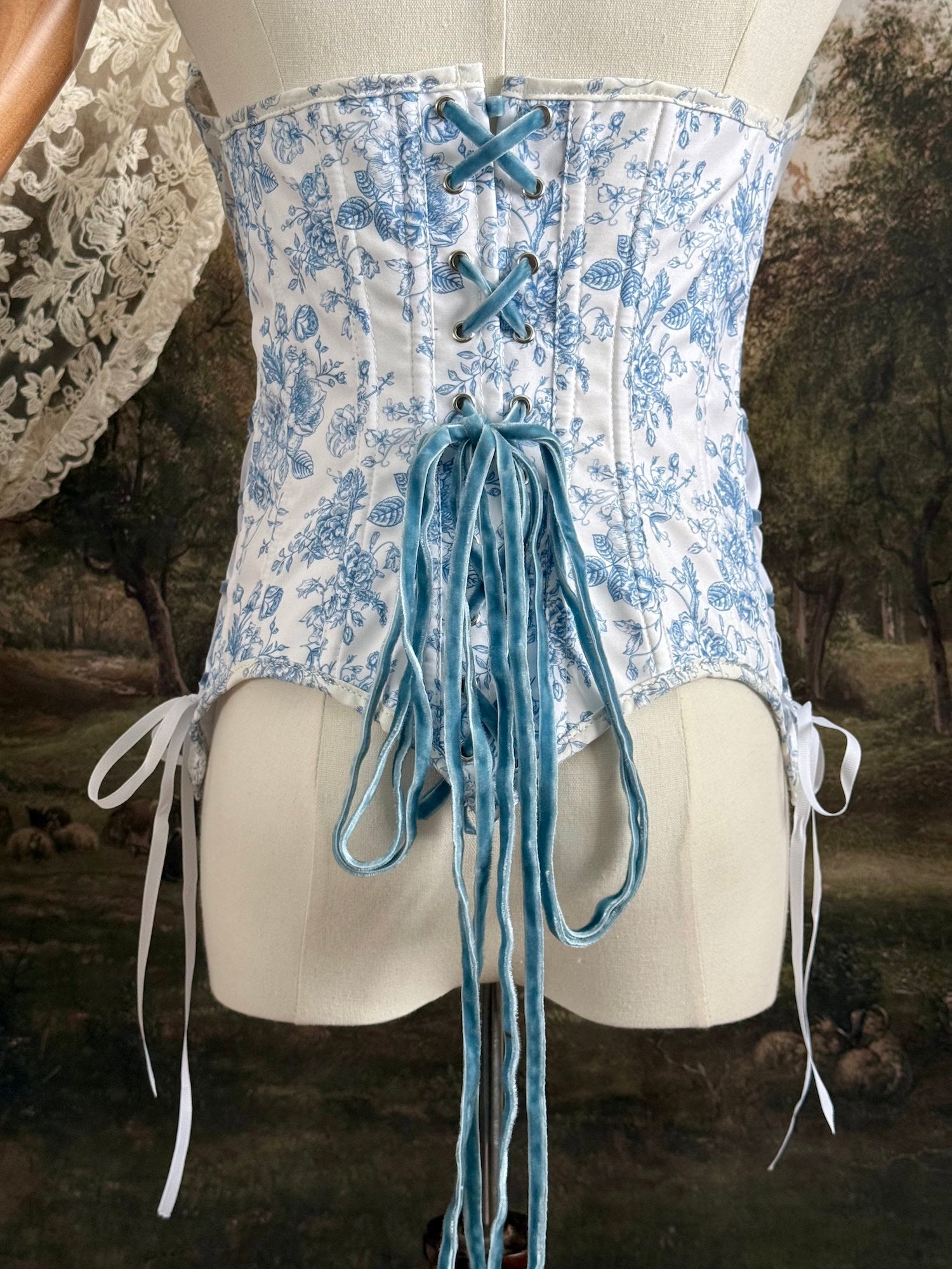 A historically-inspired vintage style overbust bustier corset in white and blue floral toile print, with a pointed basque waist, velvet lacing ties, lace trim, and a ruched bust detail, pictured on a mannequin. Back details.
