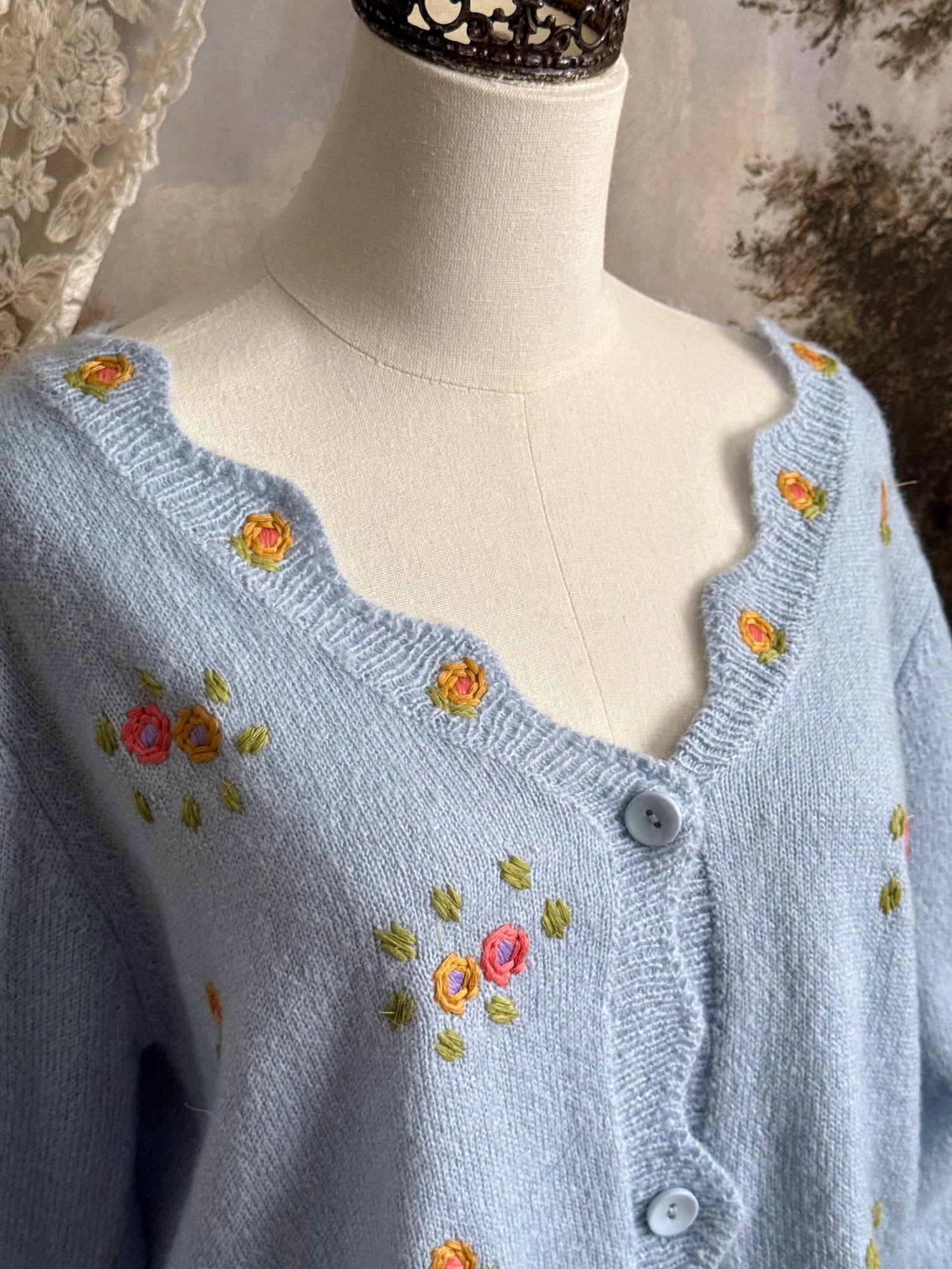 Light blue knit cardigan with vintage style floral embroidery, pictured on a mannequin against an art history background.