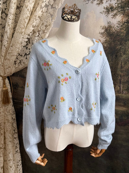 Light blue knit cardigan with vintage style floral embroidery, pictured on a mannequin against an art history background.