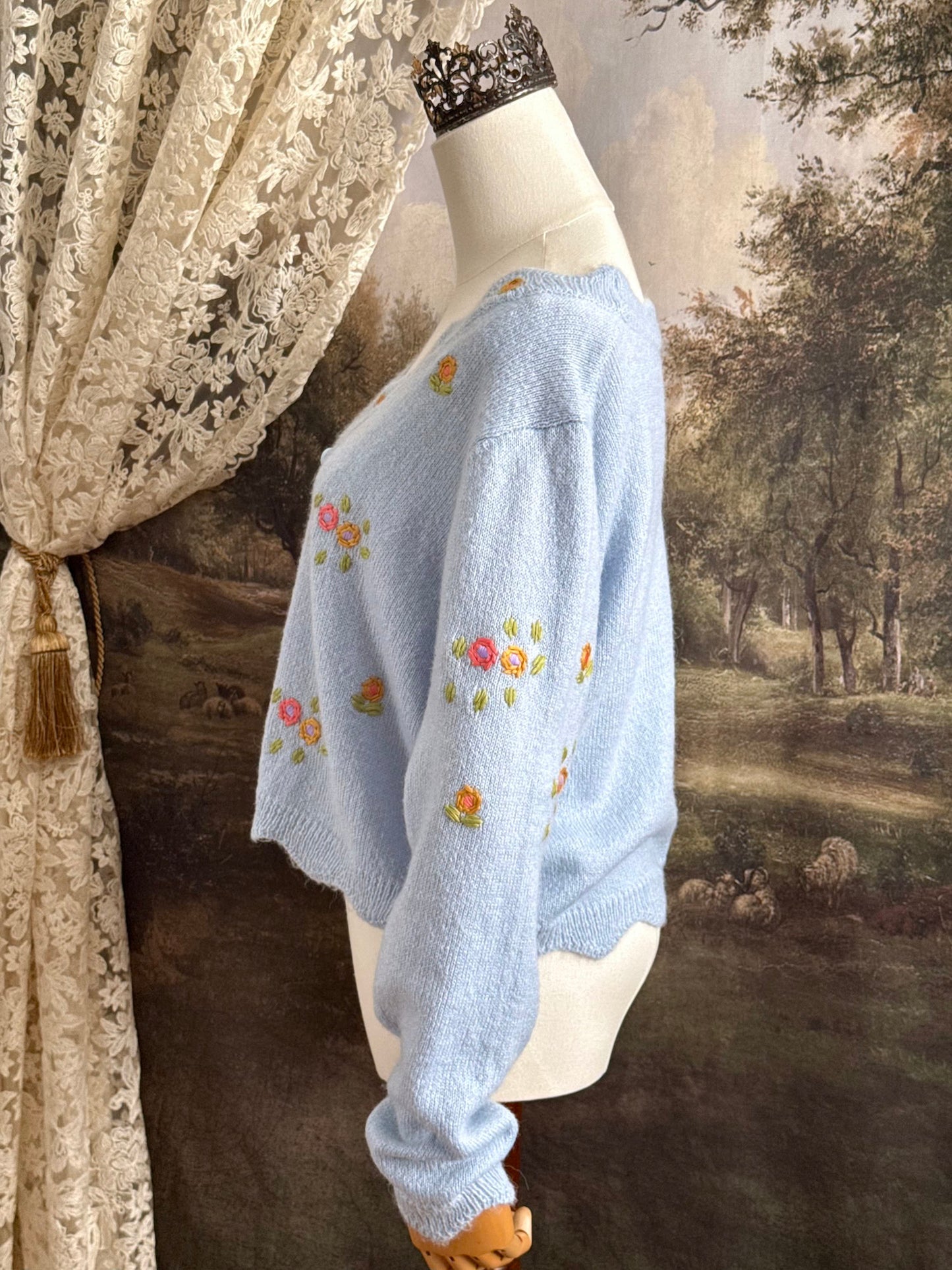 Light blue knit cardigan with vintage style floral embroidery, pictured on a mannequin against an art history background.