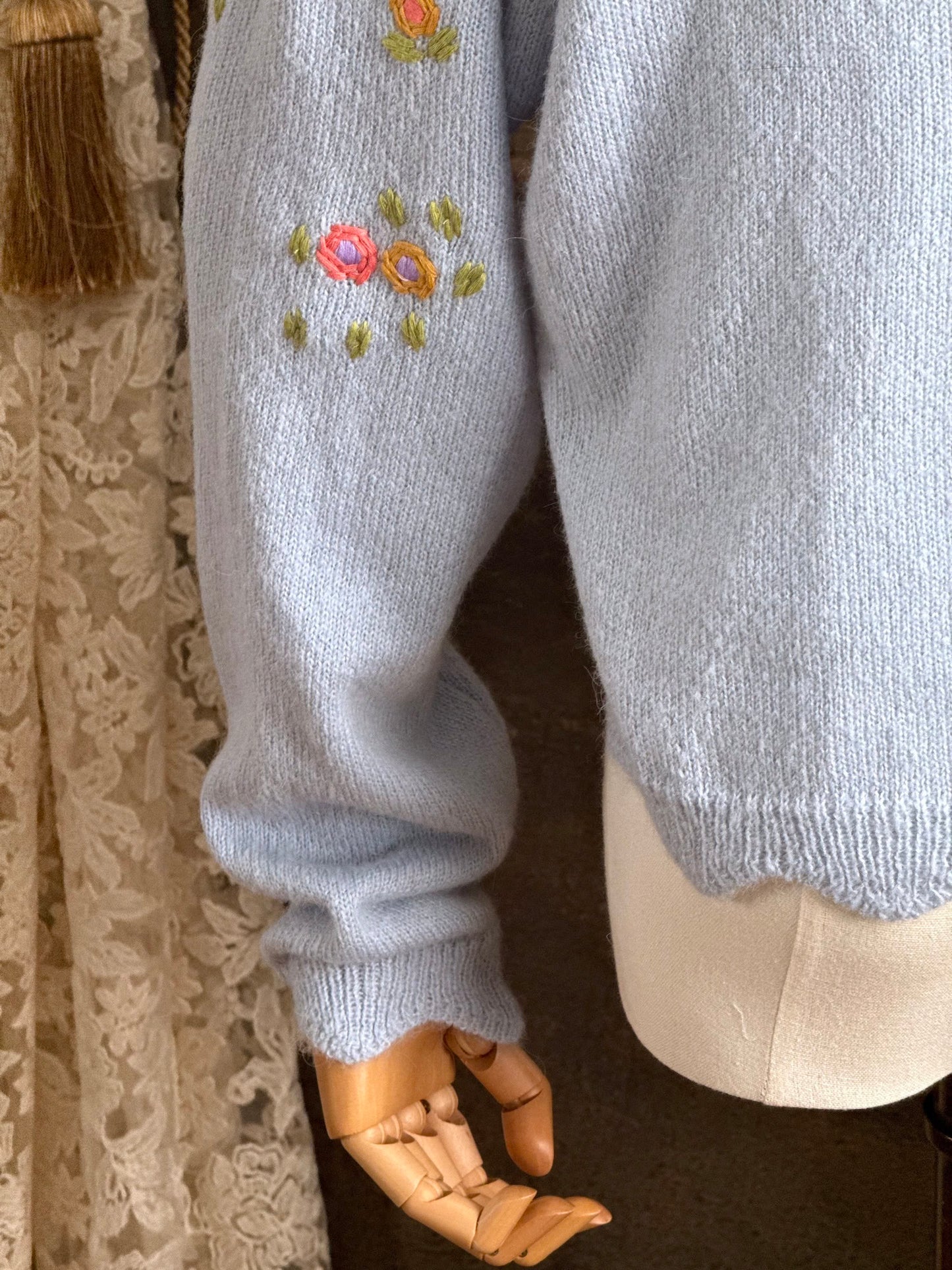 Light blue knit cardigan with vintage style floral embroidery, pictured on a mannequin against an art history background.