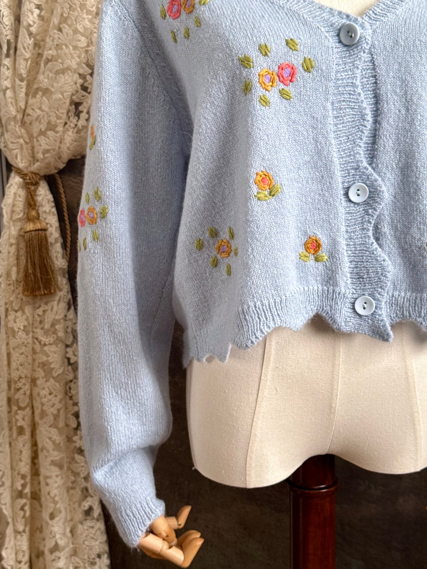 Light blue knit cardigan with vintage style floral embroidery, pictured on a mannequin against an art history background.