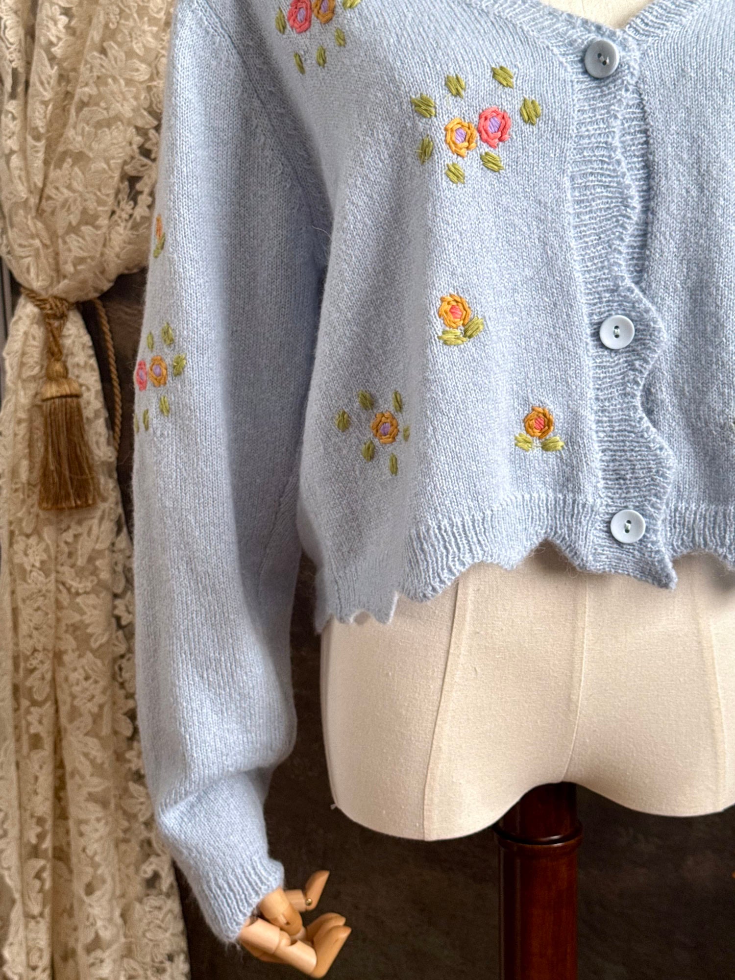 Light blue knit cardigan with vintage style floral embroidery, pictured on a mannequin against an art history background.