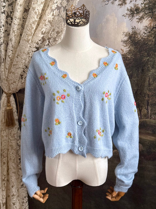 Light blue knit cardigan with vintage style floral embroidery, pictured on a mannequin against an art history background.