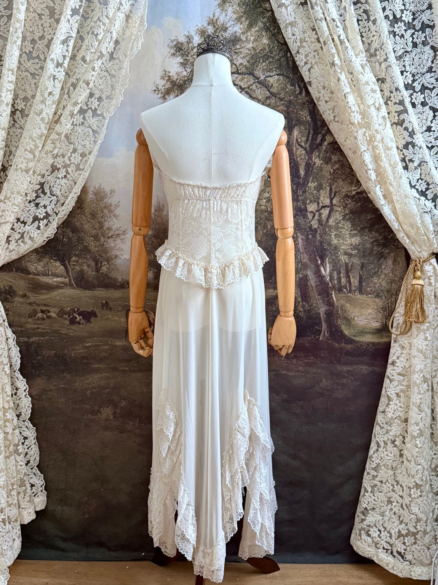 A victorian-inspired lacy chiffon overdress with ruffle lace trim and rosette appliques, pictured on a mannequin in front of a historical painted backdrop.