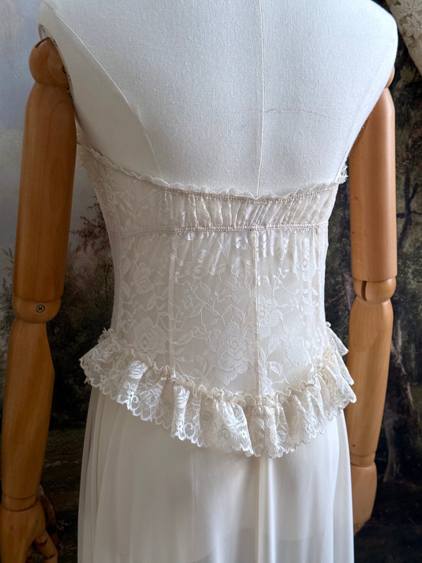 A victorian-inspired lacy chiffon overdress with ruffle lace trim and rosette appliques, pictured on a mannequin in front of a historical painted backdrop.