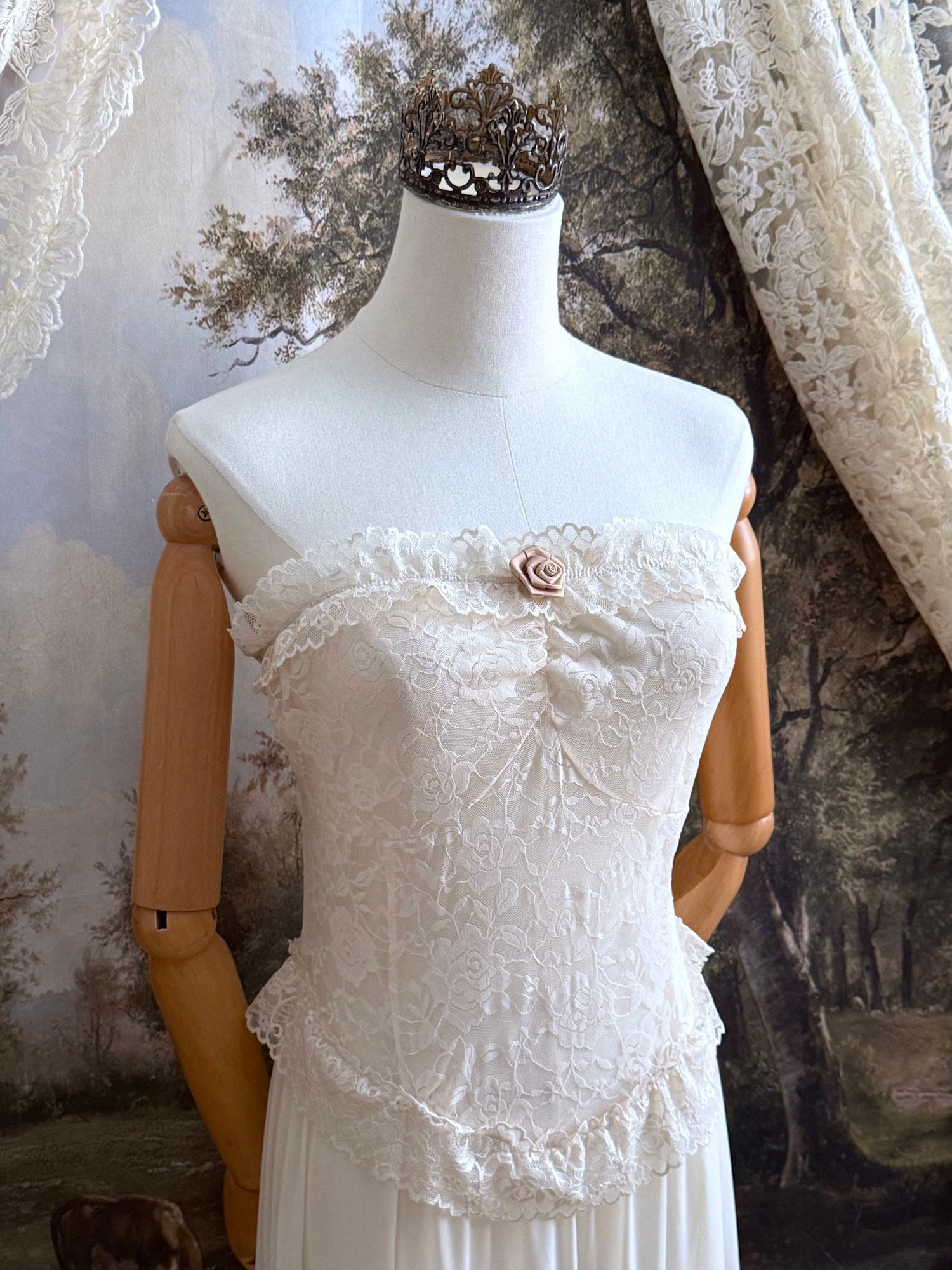 A victorian-inspired lacy chiffon overdress with ruffle lace trim and rosette appliques, pictured on a mannequin in front of a historical painted backdrop.