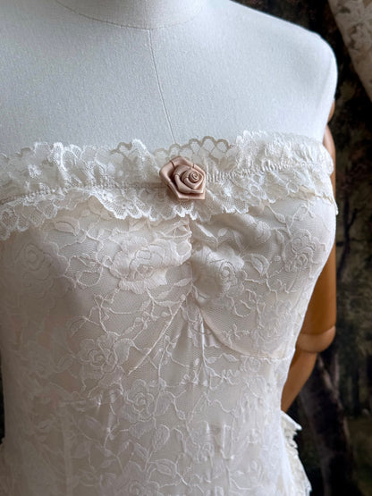 A victorian-inspired lacy chiffon overdress with ruffle lace trim and rosette appliques, pictured on a mannequin in front of a historical painted backdrop.