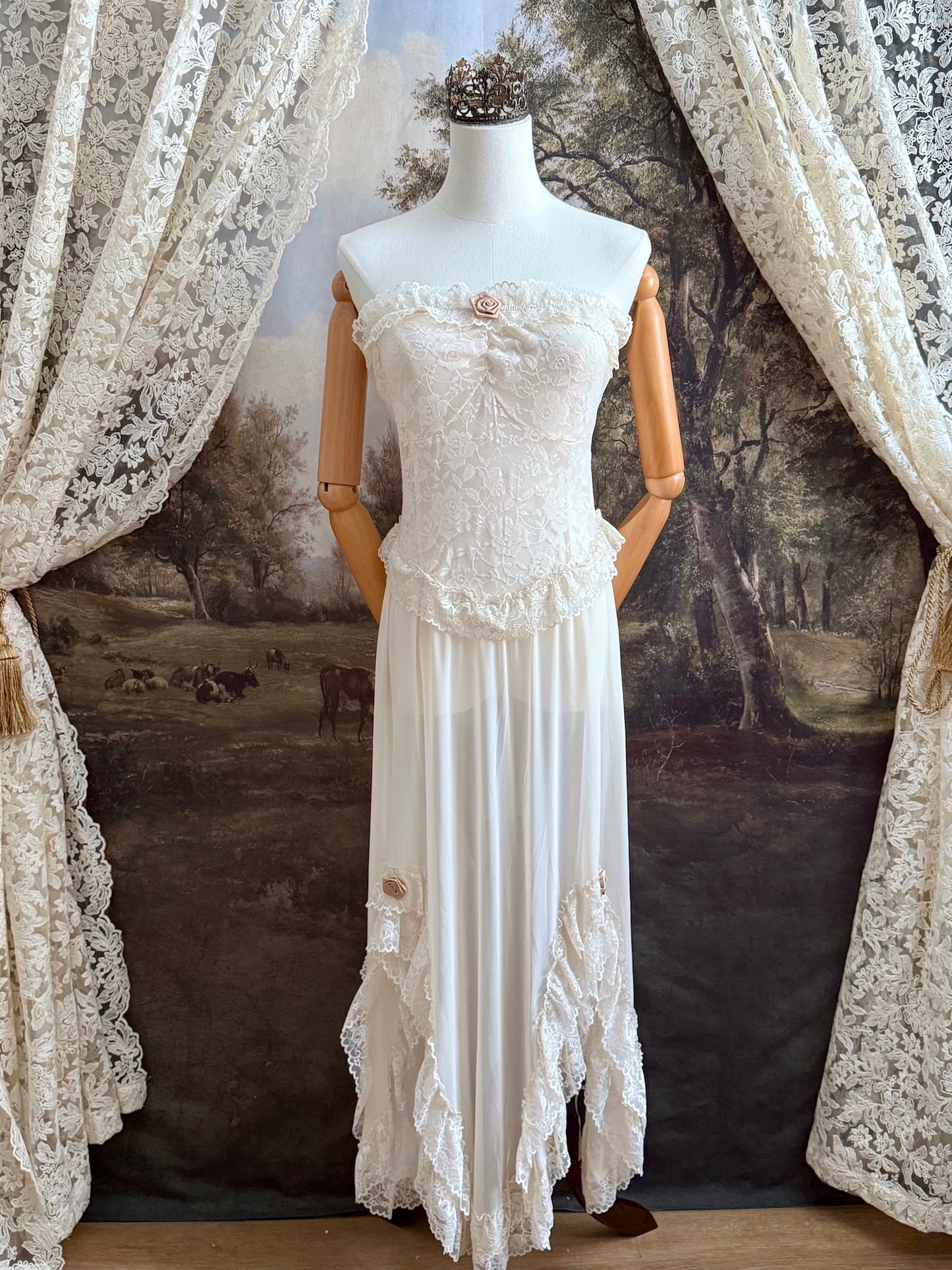 A victorian-inspired lacy chiffon overdress with ruffle lace trim and rosette appliques, pictured on a mannequin in front of a historical painted backdrop.