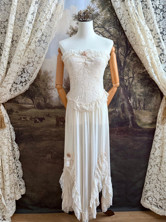 A victorian-inspired lacy chiffon overdress with ruffle lace trim and rosette appliques, pictured on a mannequin in front of a historical painted backdrop.