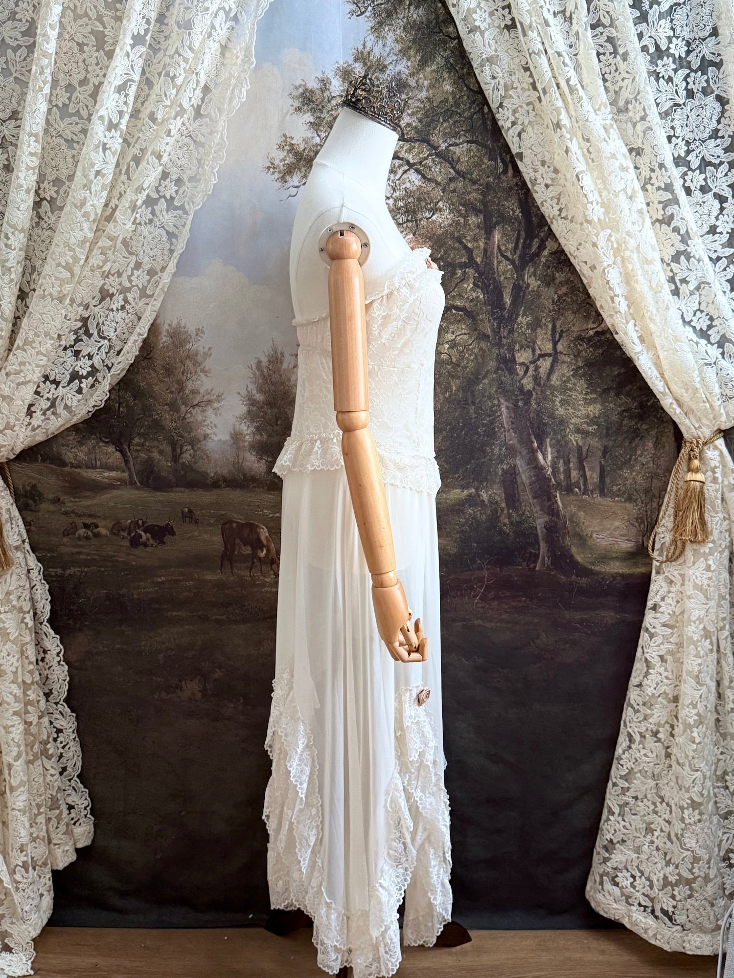 A victorian-inspired lacy chiffon overdress with ruffle lace trim and rosette appliques, pictured on a mannequin in front of a historical painted backdrop.