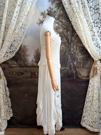 A victorian-inspired lacy chiffon overdress with ruffle lace trim and rosette appliques, pictured on a mannequin in front of a historical painted backdrop.