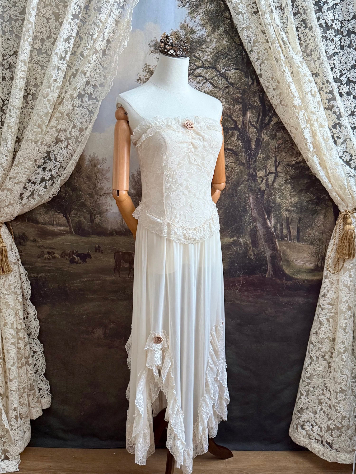 A victorian-inspired lacy chiffon overdress with ruffle lace trim and rosette appliques, pictured on a mannequin in front of a historical painted backdrop.