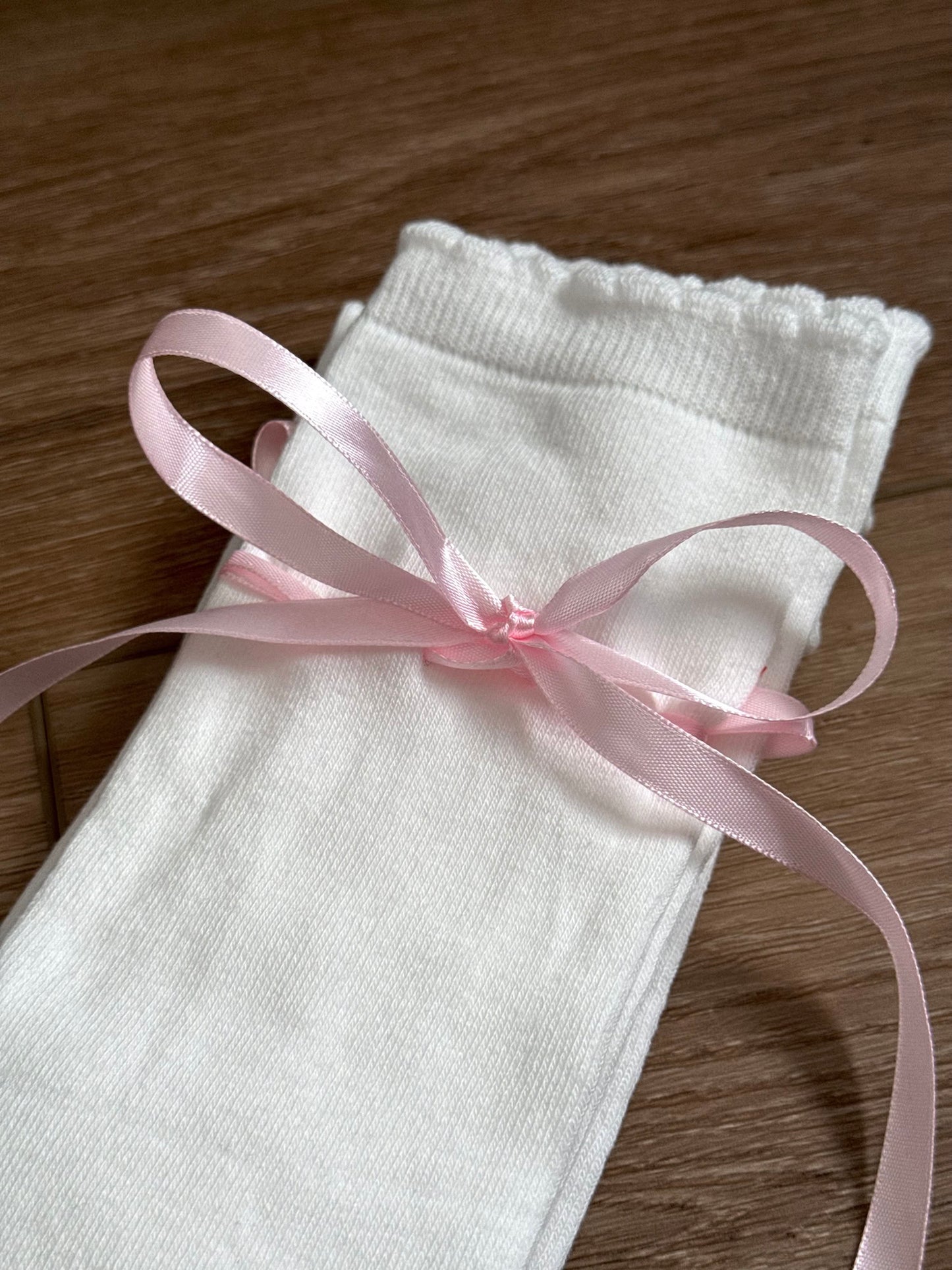 A pair of Historically Inspired Cotton Knee-High Socks with Satin Ribbon Lacing in White and Pink, pictured on a cottagecore wooden floor. For Medieval, Renaissance, Baroque, Rococo, Regency, Victorian, or Edwardian fashion. 