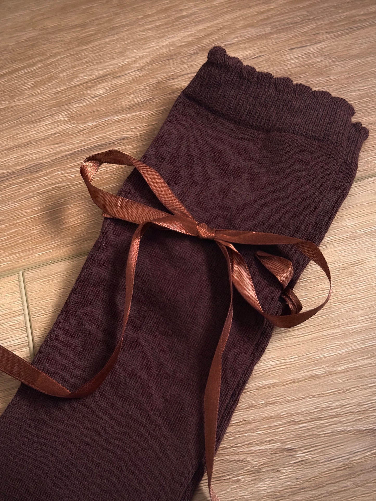 A pair of Historically Inspired Cotton Knee-High Socks with Satin Ribbon Lacing in dark brown, pictured on a historic wooden cottage floor.