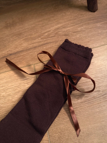 A pair of Historically Inspired Cotton Knee-High Socks with Satin Ribbon Lacing in dark brown, pictured on a historic wooden cottage floor.