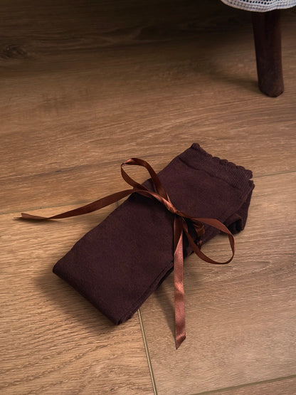 A pair of Historically Inspired Cotton Knee-High Socks with Satin Ribbon Lacing in dark brown, pictured on a historic wooden cottage floor.