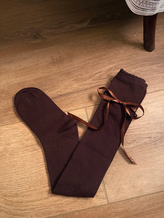 A pair of Historically Inspired Cotton Knee-High Socks with Satin Ribbon Lacing in dark brown, pictured on a historic wooden cottage floor.