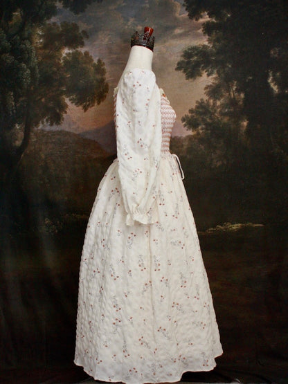 A Historically inspired rococo and regency Ivory bishop sleeve smocked tea gown with peach contrast stitching is pictured on a mannequin in front of a historical painting backdrop.