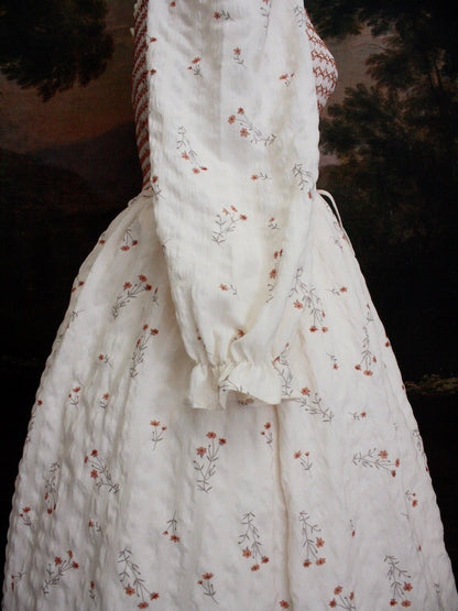 A Historically inspired rococo and regency Ivory bishop sleeve smocked tea gown with peach contrast stitching is pictured on a mannequin in front of a historical painting backdrop.